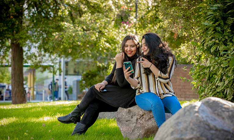 2 students sitting and chatting on a rock on campus