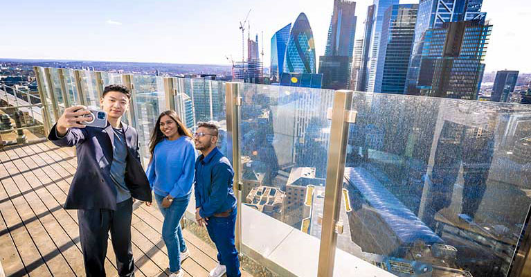 Three students on rooftop balcony taking a picture with London skyline in the background 