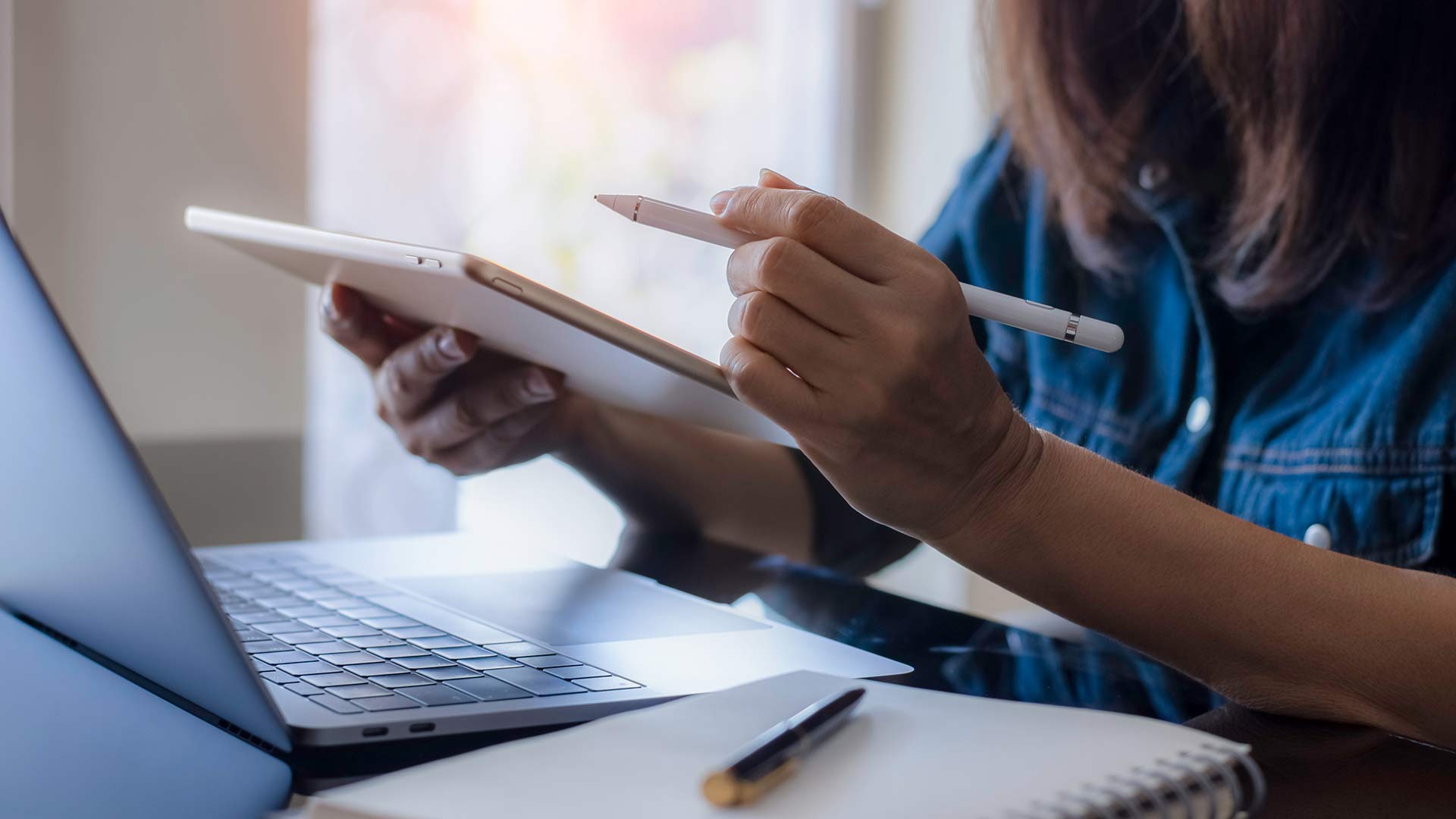 female holding a tablet with a laptop on the desk in front of her 