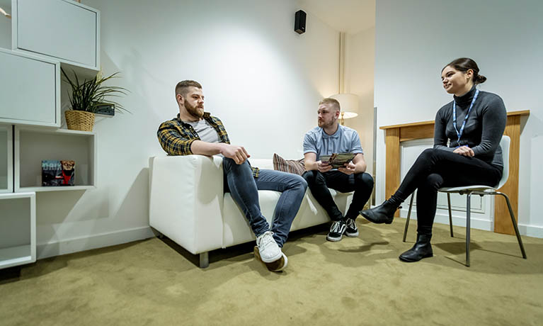 2 males sitting on a couch in conversation a female student looking on 