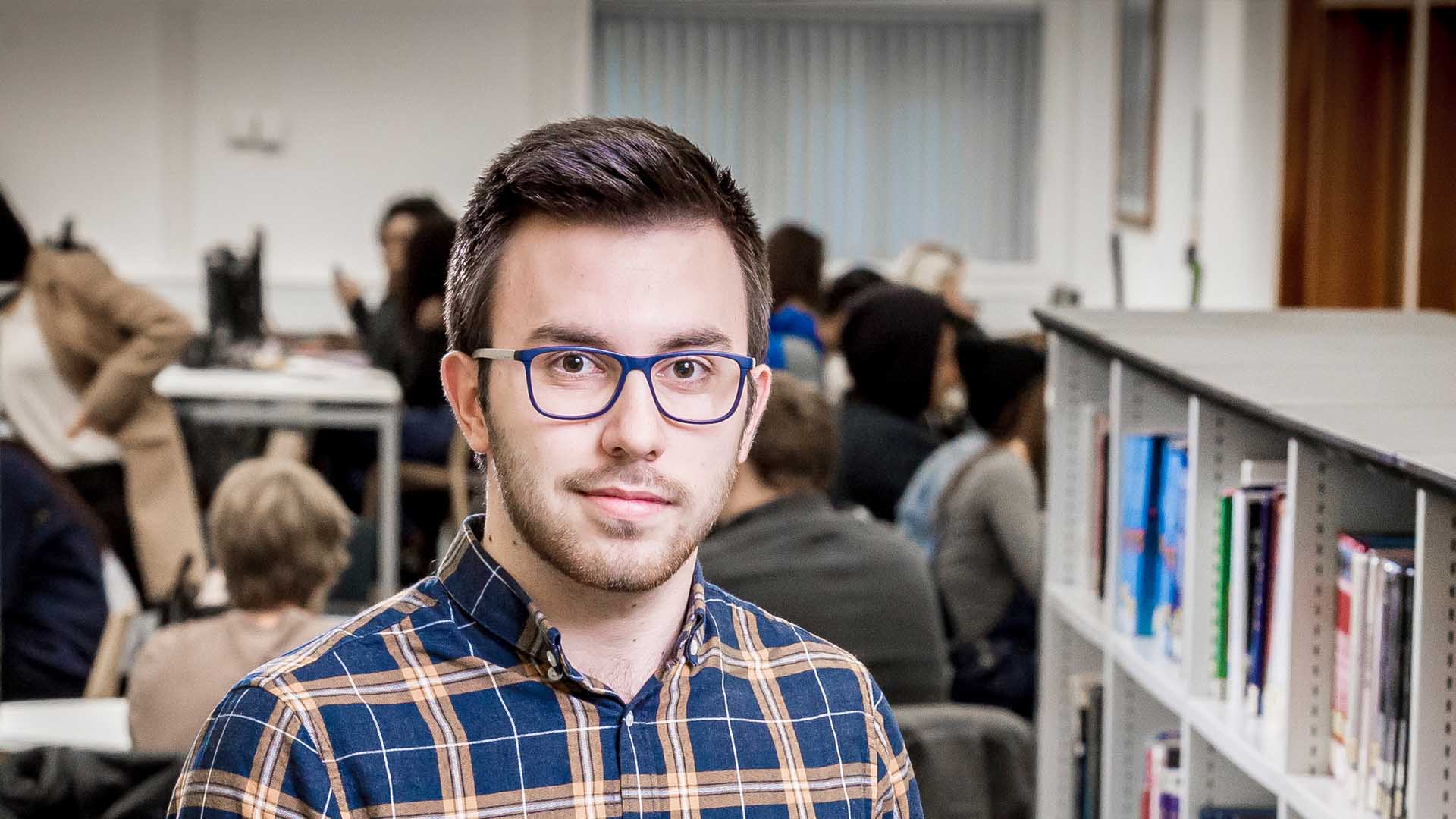 Man with glasses standing in a library next to a bookshelf