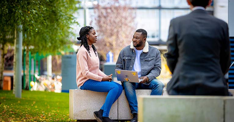 female and male sitting outside on outdoor seating. Male has an open laptop on his lap.