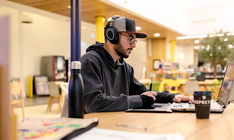 Student wearing a headphones and baseball cap working on a laptop sitting in a cafe