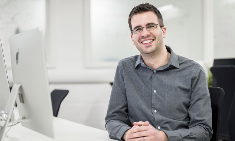 A man in a shirt behind an Apple Mac and smiling at the camera.