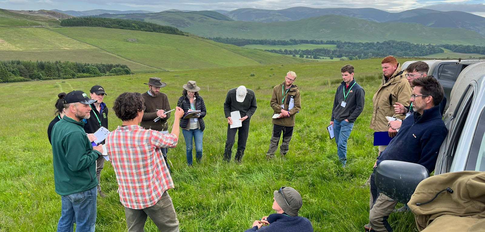 13 people gathered around in a wide circle in a field with a back drop of green hills