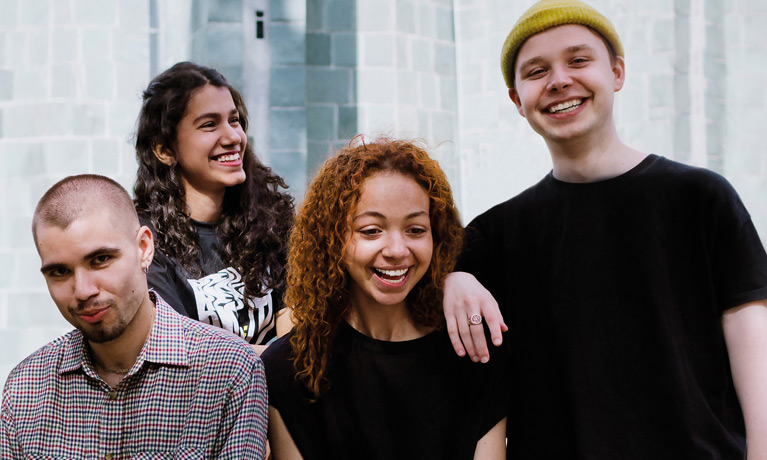 A group of four students outside a campus building, smiling at the camera.