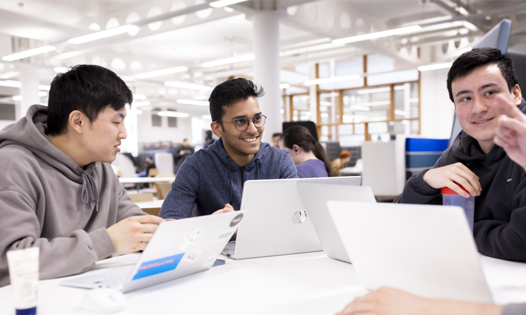 Three students talking and working on laptops in an open study space.