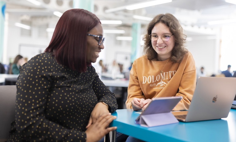 Two students in an open study area with a tablet and laptop on the table in front of them.