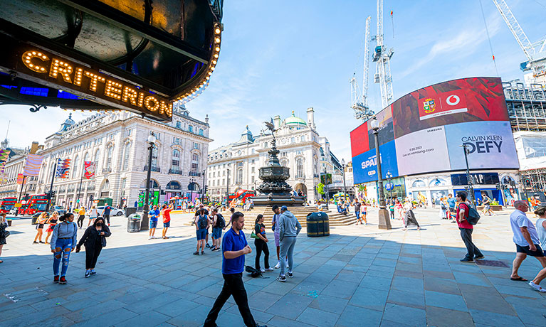 Lots of people walking across a pedestrian area in central London with lots of billboards behind them