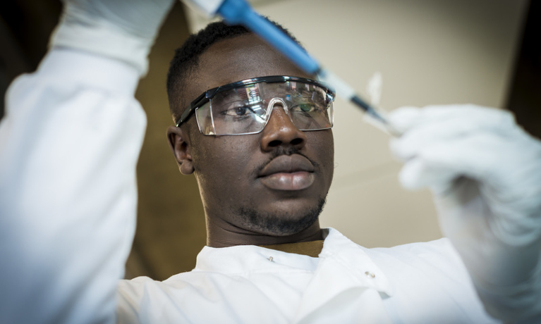 A student working in a laboratory, wearing a lab coat and protective goggles and using a pipette to transfer liquid to a test tube.