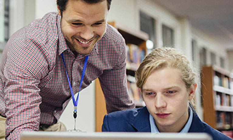 Male teacher looking at computer with pupil sat at desk.