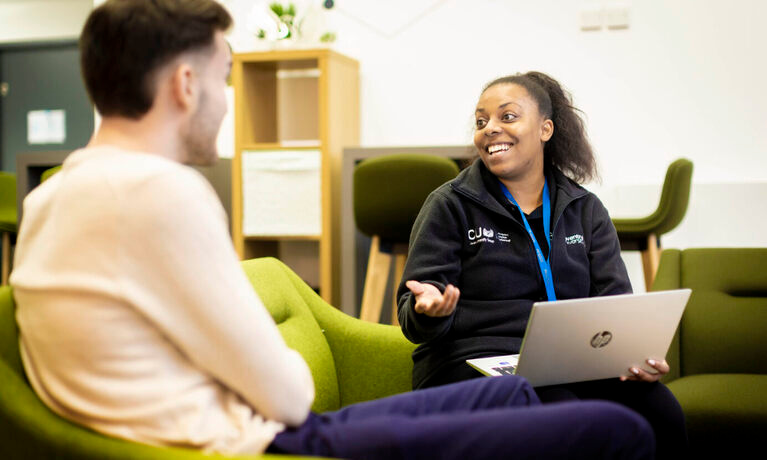 Member of staff sat down holding a laptop talking with a student